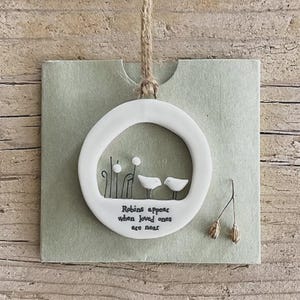 Peut inclure: Ornement circulaire en céramique blanche avec deux oiseaux et des éléments floraux. Le texte "Robins appear when loved ones are near" est inscrit. Suspendu par une corde en ficelle, sur une carte vert pâle.