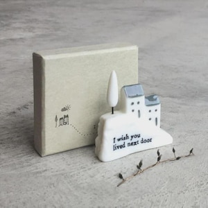 Könnte beinhalten: Eine weiße Keramikhausfigur mit einem Baum und dem Text "I wish you lived next door". Eine kleine, hellfarbene Schachtel befindet sich hinter der Figur. Ein Zweig mit Knospen liegt vor der Figur.
