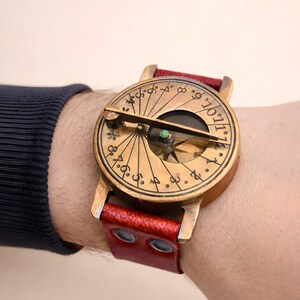 May include: A vintage brass compass watch with a red leather strap. The watch face has detailed markings and a needle. A unique accessory.