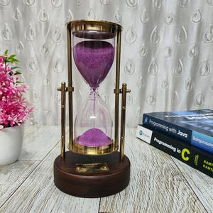 May include: An hourglass with purple sand inside, held in a clear glass container and supported by a gold-coloured metal frame. The hourglass sits on a dark wooden base with a small plaque. Books are visible in the background.