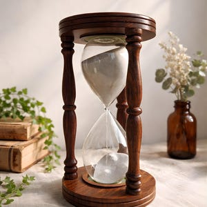 Può includere: Una clessidra in legno marrone scuro con bulbi di vetro trasparente contenenti sabbia bianca. La clessidra è sostenuta da quattro pilastri in legno tornito e una base circolare. La parte superiore e inferiore sono anch'esse circolari e in legno.
