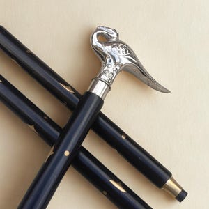 Puede incluir: Bastón negro con un mango plateado en forma de pájaro. El bastón tiene detalles dorados y una punta de latón. El mango es intrincadamente detallado y el bastón parece ser de madera.