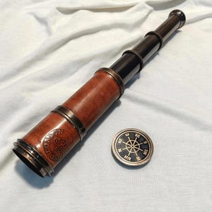 May include: A vintage-style telescope with a brown leather barrel and brass accents, alongside a small compass. The telescope has the words "LONDON GROUNDS" embossed on the side. The compass has a black face with gold lettering.