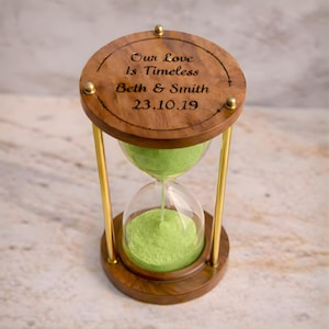 Pode incluir: Uma ampulheta de madeira com uma parte superior e inferior redondas, conectadas por hastes de metal dourado. A parte superior da ampulheta é gravada com as palavras "Our Love Is Timeless", os nomes "Beth & Smith" e a data "23.10.19". A ampulheta contém areia verde.
