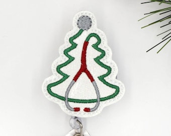 May include: A white, Christmas tree-shaped badge reel with a green outline and a red and gray stethoscope design. The badge reel has a clear plastic strap and a metal snap closure. The background is white.