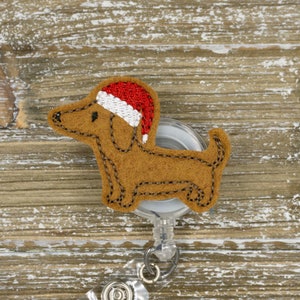 May include: A brown felt dachshund dog wearing a red and white Santa hat, attached to a clear retractable badge reel. The dog has black stitched details. The background is a light brown wooden surface.