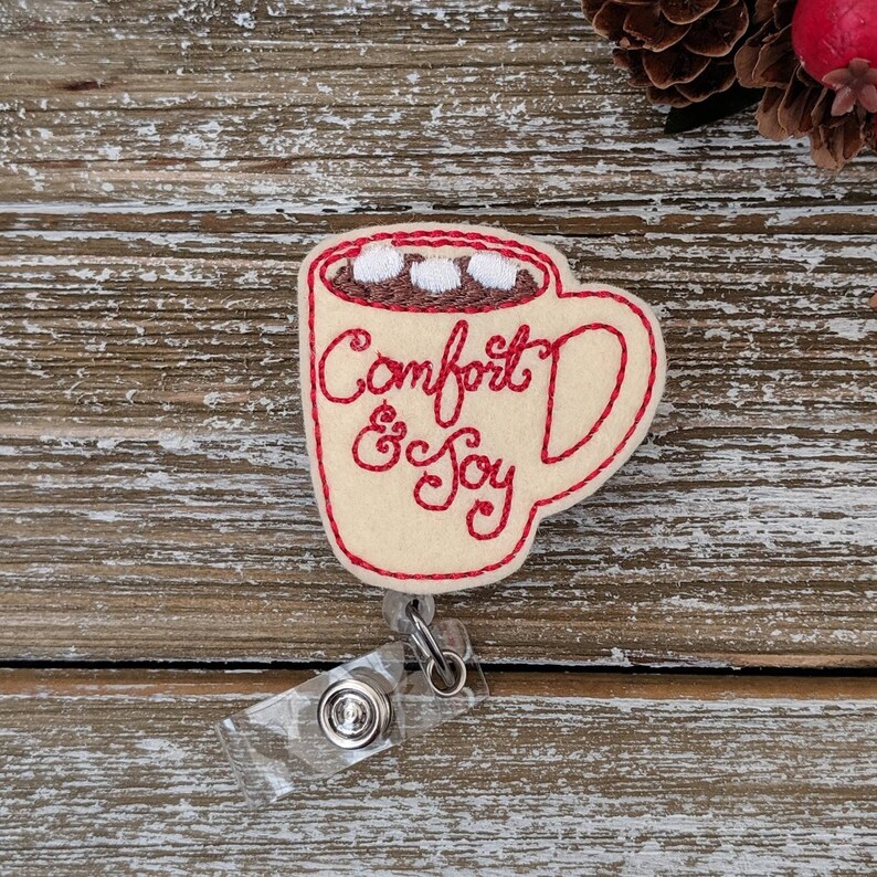 May include: A cream-colored felt badge in the shape of a mug of hot chocolate, with red stitching outlining the mug and the words "Comfort & Joy." The hot chocolate has marshmallows. A clear plastic badge holder is attached.