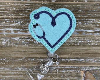 May include: A teal blue heart-shaped badge holder with a dark blue stethoscope design. The badge holder is attached to a clear retractable badge reel.