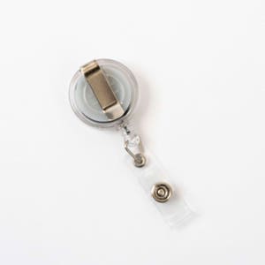 Op de afbeelding: Een doorzichtige intrekbare badgehaspel van plastic met een zilveren metalen clip en een doorzichtige plastic band met een druksluiting. De haspel heeft een grijs midden en een zilveren metalen clip.