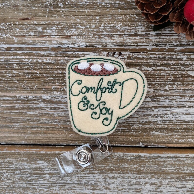 May include: A cream-colored felt badge reel in the shape of a mug of hot chocolate, with the words "Comfort & Joy" embroidered in green. The mug has a brown interior with three white marshmallows. A clear plastic reel is attached.