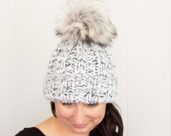 May include: A gray and white knit beanie with a large, fluffy, faux fur pom-pom on top. The hat is made of a thick, textured yarn and has a folded brim. The pom-pom is a mix of gray and white.