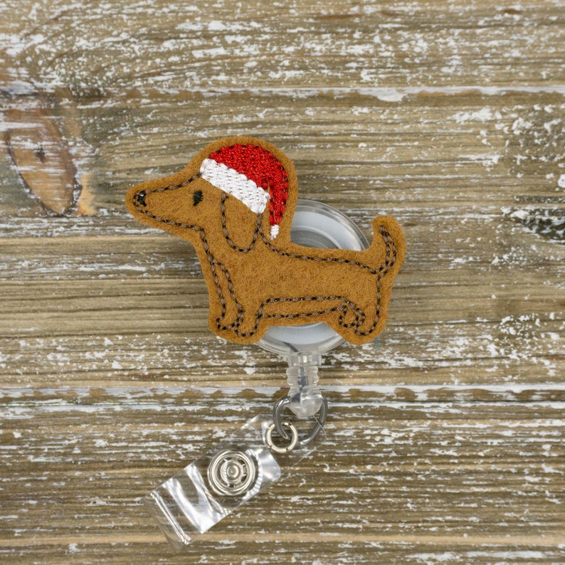 May include: A brown felt dachshund dog wearing a red and white Santa hat, attached to a clear retractable badge reel. The dog has black stitched details. The background is a light brown wooden surface.