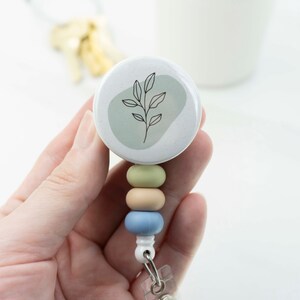 May include: A white and green badge reel with a botanical design. The reel features a round top with a leaf illustration and a pastel color bead design in green, tan, and blue. The reel is held in a hand.