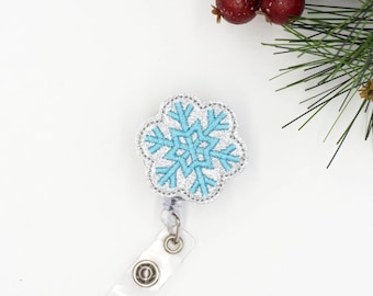 May include: A snowflake-shaped badge reel with a light blue and silver design. The badge reel has a clear plastic strap and a metal clip. The snowflake design is embroidered with a light blue center and silver outline. The background is white.
