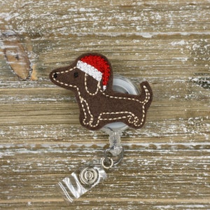 May include: A brown felt dachshund dog wearing a Santa hat, attached to a retractable badge reel. The dog has white stitched details and a red and white hat. The badge reel has a clear plastic strap and a metal clip.