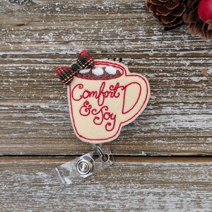 May include: A cream-colored felt badge in the shape of a mug of hot chocolate, with red embroidered text that reads "Comfort & Joy." The mug is topped with marshmallows and a plaid bow. A clear plastic badge reel is attached.