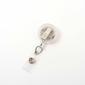 Op de afbeelding: Een heldere, ronde intrekbare badgehaspel met een metalen clip en een doorzichtige plastic band met een druksluiting. De haspel heeft een zilveren metalen clip en een doorzichtige plastic band met een druksluiting.