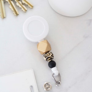 May include: A white retractable badge holder with a gold, black and white patterned bead charm. The charm features a geometric gold bead, a black and white leopard print bead, and a black bead.