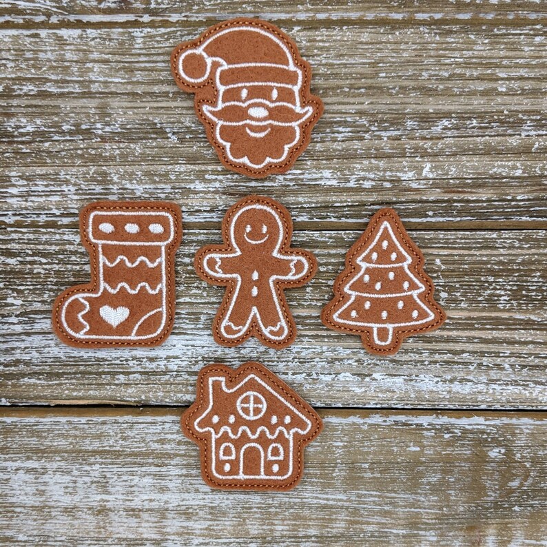 Op de afbeelding: Een verzameling bruine kerstapplicaties van vilt, waaronder de kerstman, een peperkoekmannetje, een kerstboom, een sok met een hart en een peperkoekhuisje. Elke applicatie heeft witte geborduurde details en een bruine gestikte rand.