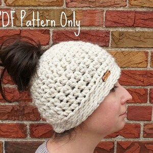 Op de afbeelding: Een witte gehaakte muts met een ruimte voor een knot aan de achterkant. De muts wordt gedragen door een persoon met bruin haar. De tekst "PDF Pattern Only" is zichtbaar bovenaan de afbeelding.