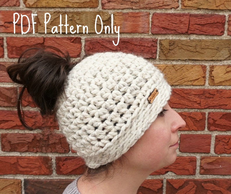 Op de afbeelding: Een witte gehaakte muts met een ruimte voor een knot aan de achterkant. De muts wordt gedragen door een persoon met bruin haar. De tekst "PDF Pattern Only" is zichtbaar bovenaan de afbeelding.
