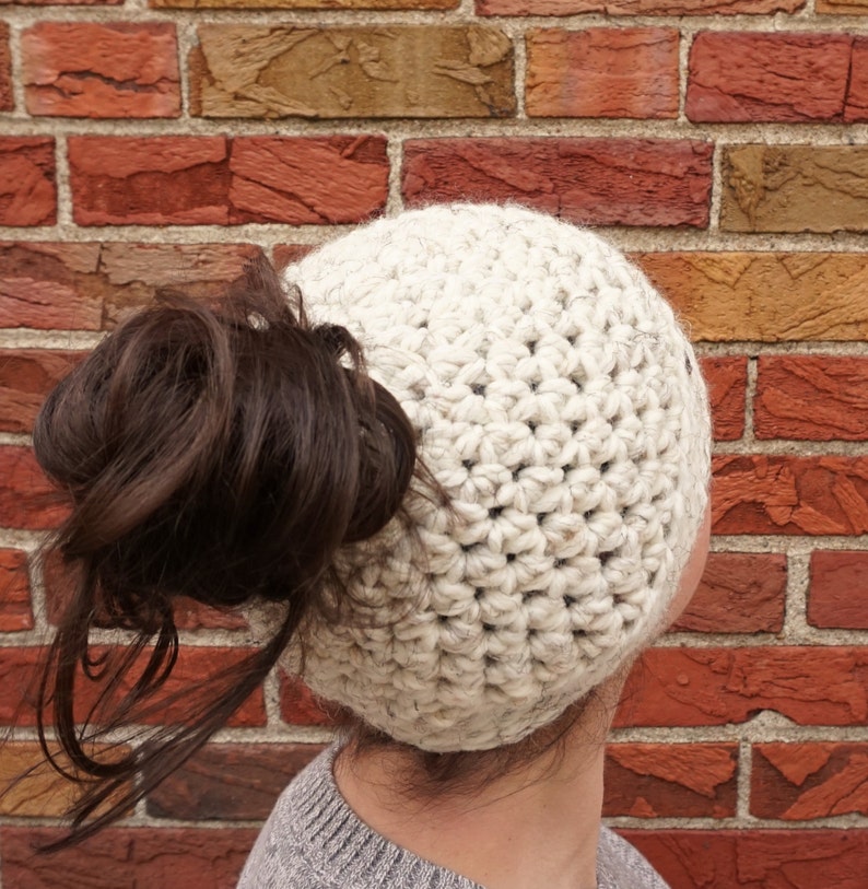 Op de afbeelding: Een witte gehaakte beanie met een gat bovenop voor een paardenstaart. De muts wordt gedragen door een persoon met bruin haar.