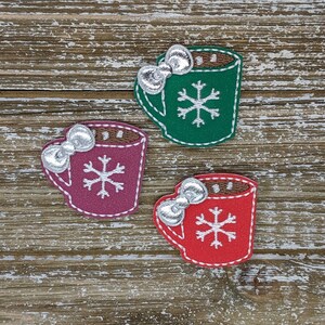 May include: Three colorful, embroidered mug-shaped patches. Each patch features a silver bow, a snowflake, and a brown rim. The mugs are in shades of green, red, and purple, with white stitching. The background is a rustic wooden surface.