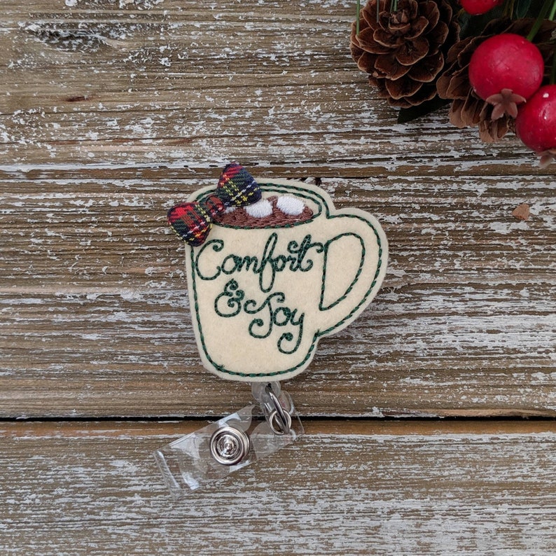 May include: A cream-colored felt badge in the shape of a mug of hot chocolate, with the words "Comfort & Joy" embroidered in green. The mug has a plaid bow and marshmallows. A clear plastic badge reel is attached.