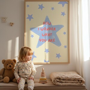 May include: A child in a floral print onesie sits on a bench next to a teddy bear and a wooden stacking toy. Above, a framed print features a light yellow background with blue stars and the text "HOW I WONDER WHAT YOU ARE" in red and blue.