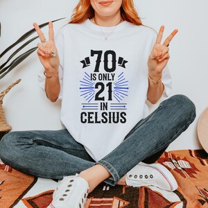 以下が含まれることがあります： 白地に「70 is only 21 in Celsius」の文字が黒と青でプリントされたTシャツ。カジュアルなスタイルで、普段使いに適しています。年齢を遊び心で表現したデザインです。