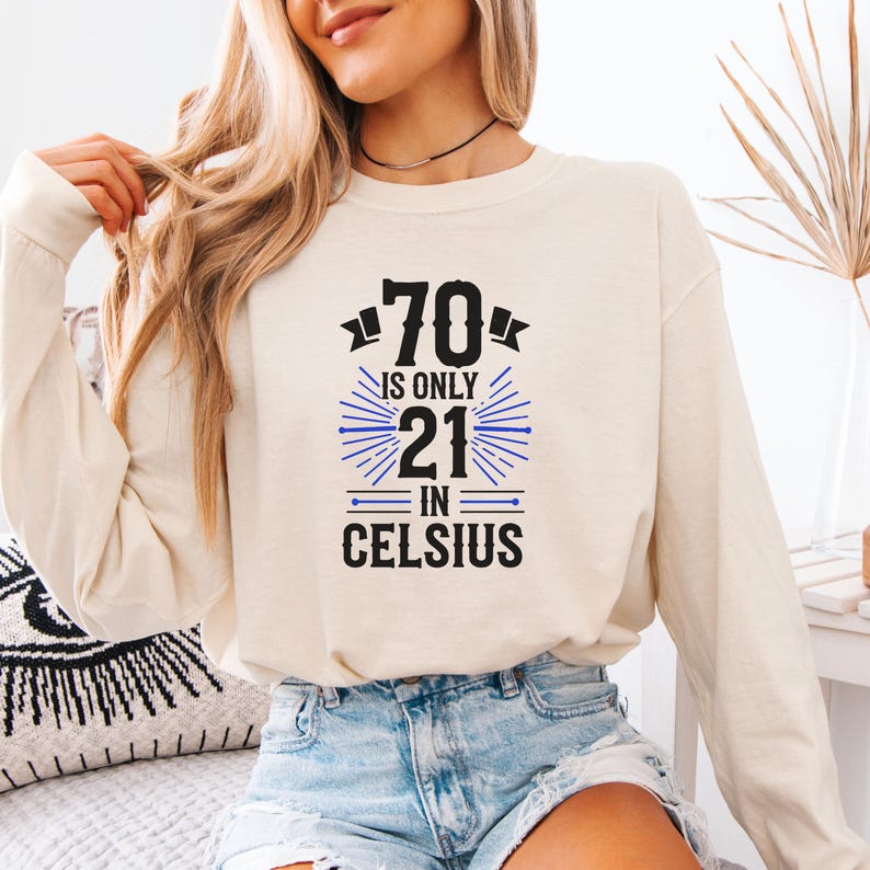 以下が含まれることがあります： 「70 is only 21 in Celsius」のテキストが黒と青でプリントされた、クリーム色の長袖シャツ。金髪とジーンズを着用した人が着ています。