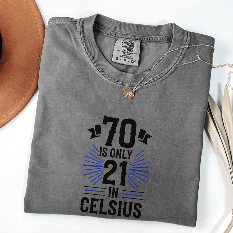 以下が含まれることがあります： 黒と青で「70 is only 21 in Celsius」と書かれたグレーのComfort Colors Tシャツ。ペンダント付きのシルバーネックレスがシャツの上にかけられています。シャツは白い面に折りたたまれています。