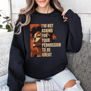 Puede incluir: Sudadera azul marino oscuro con un gráfico de un perfil de mujer en tonos naranja y marrón, y el texto "I'M NOT ASKING FOR YOUR PERMISSION TO BE GREAT".