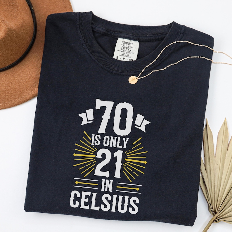 以下が含まれることがあります： 「70 is only 21 in Celsius」の文字が白と黄色でプリントされたネイビーブルーのTシャツ。クルーネックで、Comfort Colorsのタグが付いています。ゴールドのネックレスと茶色の帽子も写っています。