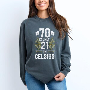 以下が含まれることがあります： 白と黄色の「70 is only 21 in Celsius」の文字が入った、濃いグレーの長袖スウェットシャツ。 グラフィックには数字の70、数字の21、そしてCelsiusという単語が描かれています。
