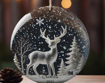 May include: A clear, round Christmas ornament with a winter scene. Inside, a white deer stands in a snowy landscape with trees. The ornament has the name "Charlotte" and the year "2025" printed on it. The ornament hangs from a string.