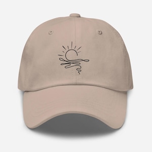 May include: A beige baseball cap featuring a black line art graphic on the front panel. The design depicts a stylized sun rising or setting over wavy water, rendered in a single continuous black line. The cap has a curved brim and eyelets.