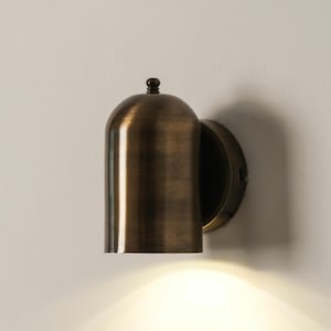 Op de afbeelding: Een bronzen wandlamp met een klokvormige kap en een ronde montageplaat. Het licht schijnt naar beneden en verlicht de ruimte eronder. De armatuur heeft een klassiek, vintage ontwerp.