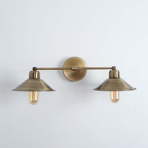 May include: A two-light wall sconce in a vintage style. The brass-finished fixture features two conical shades and exposed bulbs. It is mounted on a round backplate and a horizontal bar. The design evokes industrial or farmhouse aesthetics, suitable for various interiors.