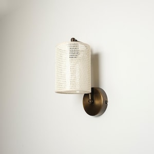 May include: A wall-mounted light fixture with a cylindrical, off-white woven shade. The fixture has a dark bronze-colored base and mounting hardware. The shade is a textured, open weave, allowing light to filter through. The design is modern and minimalist.