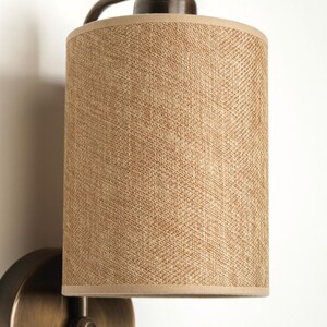 May include: A wall-mounted light fixture with a cylindrical lampshade. The lampshade is made of a woven, tan-colored material with a light brown trim. The fixture has a dark bronze finish.