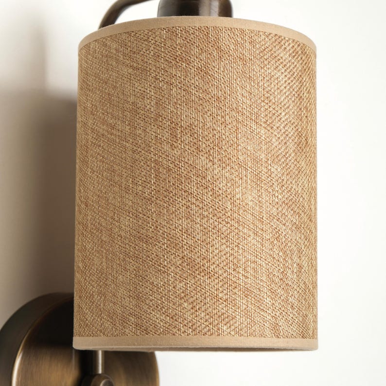 May include: A wall-mounted light fixture with a cylindrical lampshade. The lampshade is made of a woven, tan-colored material with a light brown trim. The fixture has a dark bronze finish.