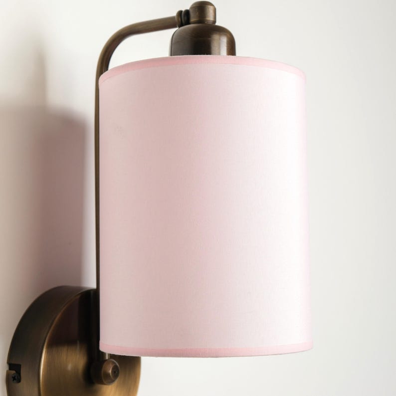 May include: A wall-mounted lamp with a cylindrical pink lampshade and a bronze-colored metal frame. The lampshade has a light pink trim. The lamp is designed to be mounted on a wall and provides ambient lighting.