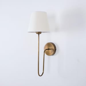 Op de afbeelding: Een wandlamp met een crèmekleurige lampenkap en een messingkleurig metalen frame. De lamp heeft een gebogen arm die de kap verbindt met een ronde wandbevestiging. Het ontwerp is eenvoudig en elegant.