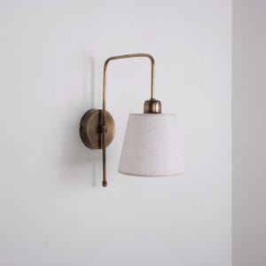 Op de afbeelding: Een messing wandlamp met een witte lampenkap. De lamp heeft een gebogen arm en een conische kap. Deze lamp is ontworpen voor binnenverlichting en voegt een vleugje vintage stijl toe.