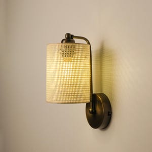 May include: A wall-mounted light fixture with a cylindrical shade made of a woven, light-colored material. The fixture has a bronze-colored metal frame and a round base. The light is on, illuminating the shade.