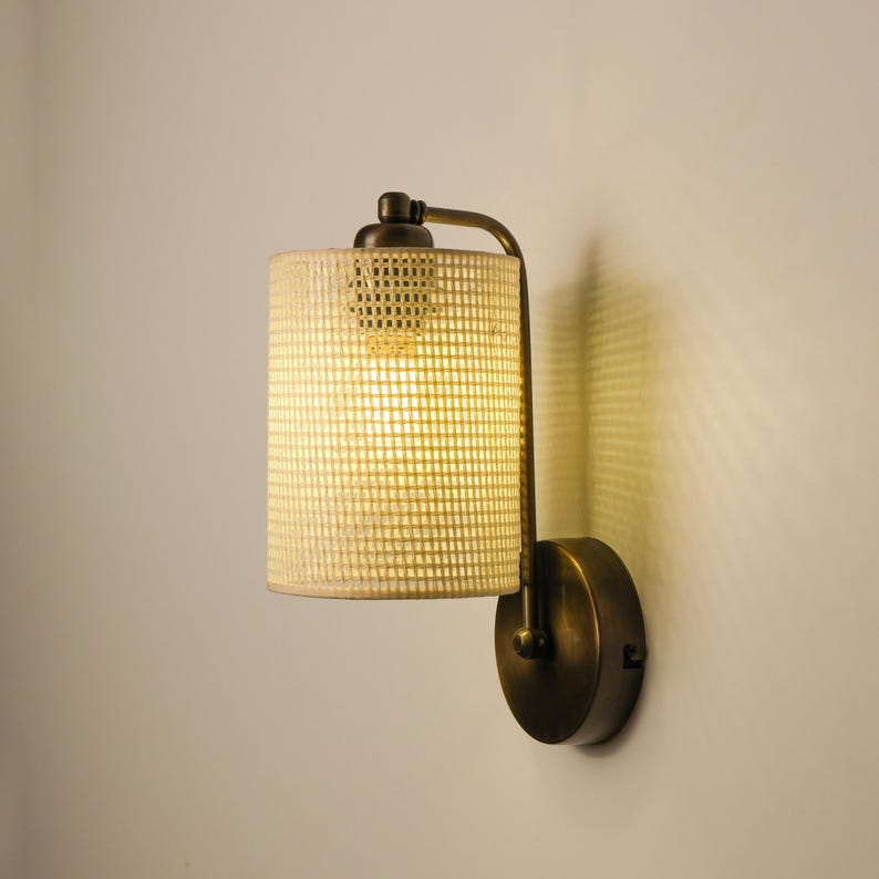May include: A wall-mounted light fixture with a cylindrical shade made of a woven, light-colored material. The fixture has a bronze-colored metal frame and a round base. The light is on, illuminating the shade.