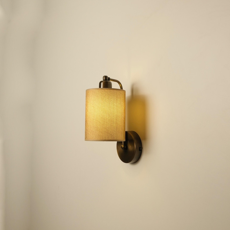 May include: A wall-mounted sconce with a cylindrical lampshade. The shade is a light tan color, and the fixture has a dark bronze finish. The sconce is illuminated, casting a warm glow.