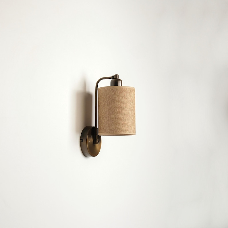 May include: A wall-mounted sconce with a cylindrical beige lampshade. The fixture has a dark bronze finish, with a curved arm connecting the shade to a circular base. The lamp is designed for indoor use and provides ambient lighting.