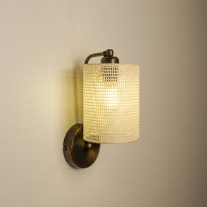 May include: A wall-mounted light fixture with a cylindrical, off-white shade featuring a grid pattern. The fixture has a bronze-colored base and arm, and the light is illuminated. The design is simple and modern.